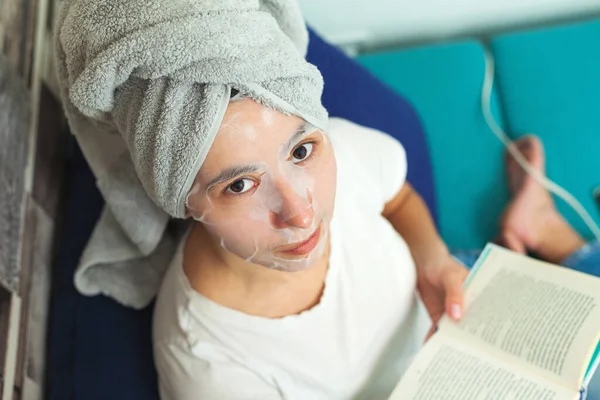 Girl in a cosmetic mask and a towel on her head. Young woman reads a book and does cosmetic procedures.