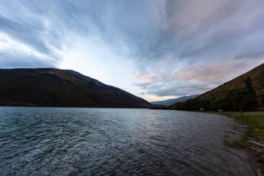 Aydınlatılmış tepeleri olan yüksek kayalıkları olan güzel bir manzara, dağ gölündeki taşlar, yansıma, mavi gökyüzü ve gündoğumunda sarı güneş ışığı. 