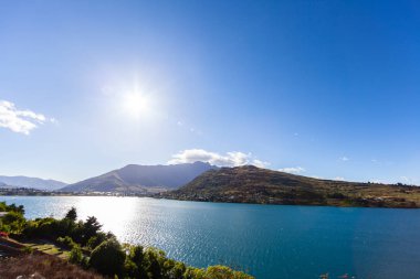 Aydınlatılmış tepeleri olan yüksek kayaları olan güzel bir manzara, dağ gölündeki taşlar, yansıma, mavi gökyüzü ve gün doğumunda sarı güneş ışığı. Tekapo, Queenstown, Wanaka.