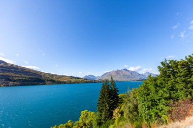Aydınlatılmış tepeleri olan yüksek kayaları olan güzel bir manzara, dağ gölündeki taşlar, yansıma, mavi gökyüzü ve gün doğumunda sarı güneş ışığı. Tekapo, Queenstown, Wanaka.