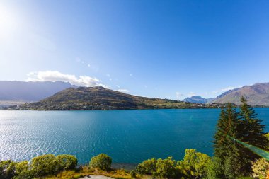 Aydınlatılmış tepeleri olan yüksek kayaları olan güzel bir manzara, dağ gölündeki taşlar, yansıma, mavi gökyüzü ve gün doğumunda sarı güneş ışığı. Tekapo, Queenstown, Wanaka.