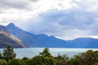 Aydınlatılmış tepeleri olan yüksek kayaları olan güzel bir manzara, dağ gölündeki taşlar, yansıma, mavi gökyüzü ve gün doğumunda sarı güneş ışığı. Tekapo, Queenstown, Wanaka.