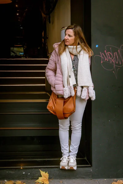 White woman with pants and coat leaning against the wall and loo