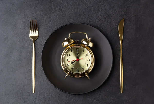 Arany ébresztőóra egy fekete tányéron arany késsel és villával — Stock Fotó