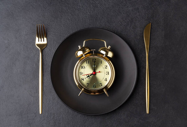 Golden alarm clock on a black plate with a golden knife and fork
