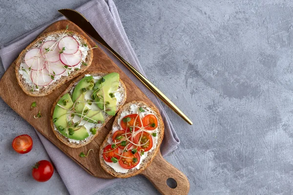 Три бутерброда на разделочной доске с зерновым хлебом, овощами и микрозеленью на сером фоне — стоковое фото