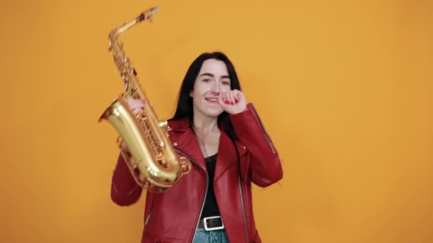 Šílená usmívající se mladá žena drží saxofon, ukazuje dlaně přes oranžovou zeď — Stock video