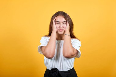 Yorgun genç bir kadın ellerini kafasından ayırmıyor, baş ağrısı turuncu duvarda izole edilmiş.