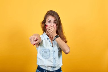 Şok olmuş, beyaz bir genç kadın. Elini ağzına koyup kamerayı işaret ediyor.