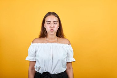 Çekici bir genç kadın, yanaklarında hava tutuyor, turuncu duvarda ıssız bir şekilde esiyor.