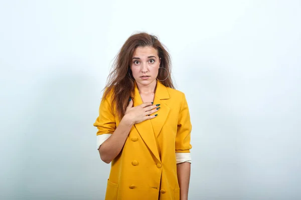 Šokovaná mladá žena ve žluté bundě drží ruce na prsou, vypadá děsivě — Stock fotografie