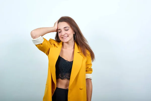 Aantrekkelijke jonge vrouw in geel jasje houdt hand op hoofd, glimlachend — Stockfoto