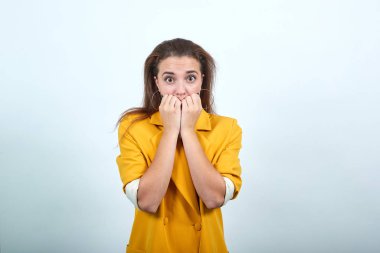 Scared young woman in yellow jacket keeping hand on mouth, biting her nail