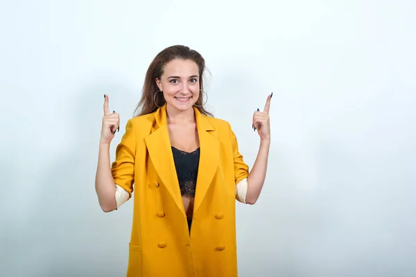 Привлекательная юная леди в желтой куртке, показывающая пальцем вверх, выглядящая счастливой — стоковое фото