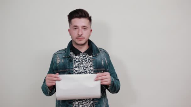 Jonge man in de mode denim jas houden mooi wit papier, op zoek lachend — Stockvideo