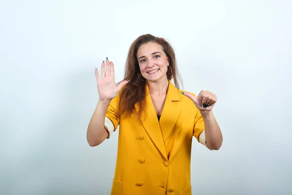 Atrakcyjna młoda kobieta pokazująca sześć z palcem, wyglądająca szczęśliwie w żółtej kurtce — Zdjęcie stockowe