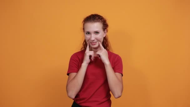 Bonita joven de camisa roja con la mano en las mejillas, sonriendo — Vídeo de stock