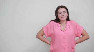 Pembe gömlekli ciddi bir kadın ellerini başının üstünde tutuyor, diğer elini arkada tutuyor.