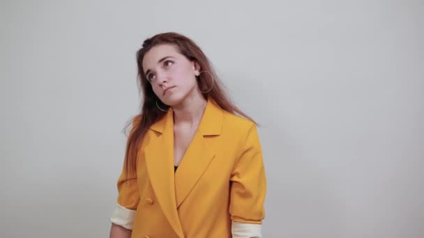 Mujer joven bonita en chaqueta amarilla manteniendo la mano en la mejilla, buscando aburrido — Vídeo de stock