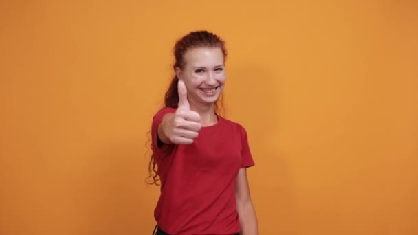Усміхнена молода жінка в червоній сорочці вказує напряму, посміхаючись над помаранчевою стіною — стокове відео