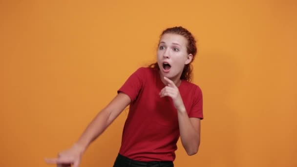 Bang jonge vrouw in rood shirt wijzend in de verte, geïsoleerd op oranje muur — Stockvideo