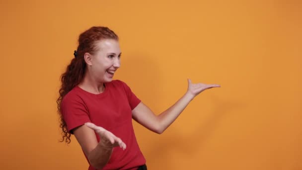 Привлекательная юная леди в красной рубашке приветствует, приглашает кого-то через оранжевую стену — стоковое видео