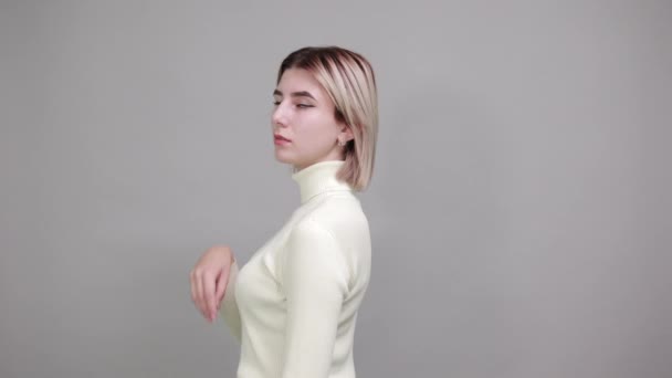Mujer joven estricta en suéter blanco, cogida de la mano en la barbilla sobre la pared gris — Vídeos de Stock