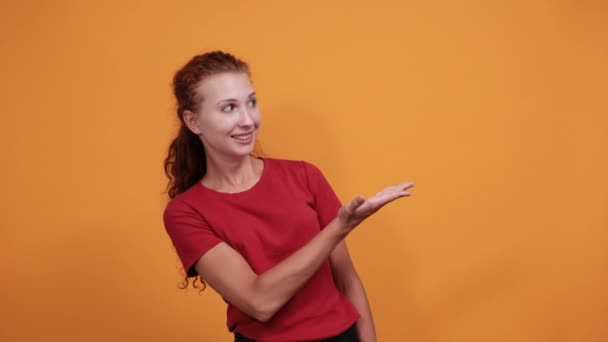 Bonita joven con camisa roja mostrando espacio a un lado, sonriendo, luciendo feliz — Vídeo de stock