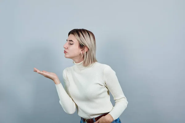 Söt ung kvinna i vit tröja skicka luft kyss åt sidan isolerad på grå vägg — Stockfoto