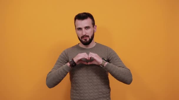 Серйозний красивий кавказький чоловік робить форму серця, виглядає красиво, посміхаючись — стокове відео