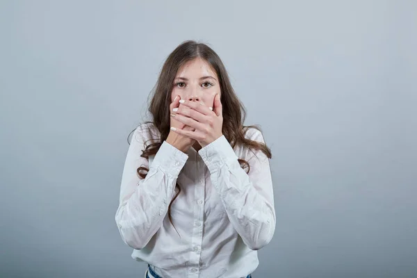 Scared young woman in white shirt covering mouth with hands, looking shocked — 스톡 사진
