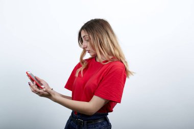 Moda kırmızısı tişörtlü çekici bir kadın telefonu elinde tutuyor ve ona bakıyor.