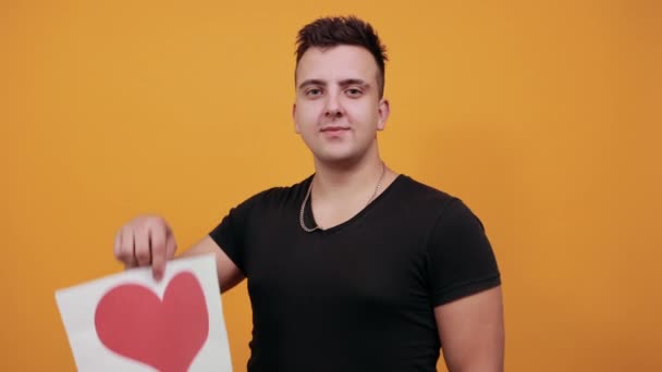 Cheerful young man keeping picture with red heart, showing victory symbol. — 비디오