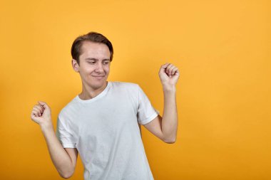 Sarı arka planda beyaz tişörtlü genç adam mutluluktan ellerini kaldırıyor..