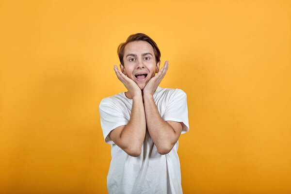 Молодой человек с удивленной гримасой поддерживает подбородок руками в белой футболке
