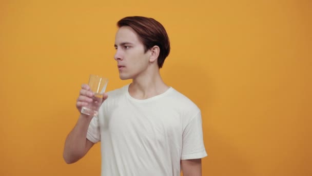 Een man in een wit t-shirt op een gele achtergrond drinkwater uit een glas — Stockvideo