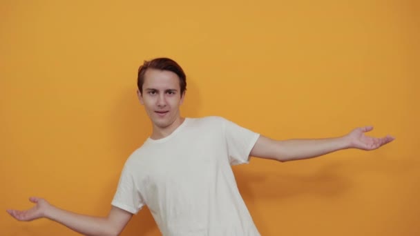 Handsome young man shows his hands wide. In white t-shirt on yellow background — Stock Video