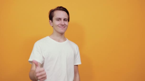 Um jovem de t-shirt branca no fundo amarelo dá polegares para cima — Vídeo de Stock