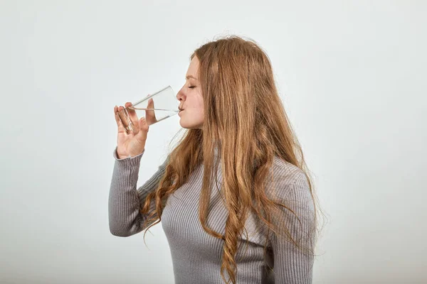Mulher de cabelos vermelhos em camisola cinza sacia sua sede e bebe copo de água — Fotografia de Stock