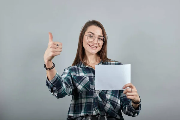happy woman with glasses holds a white sheet of paper in hand, shows her thumb