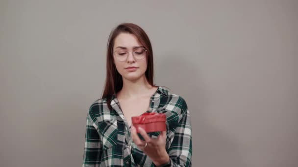 Upset woman in glasses holds gift box in shape of heart, touches finger tongue — 图库视频影像