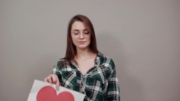 Beledigde vrouw in bril houdt een geschenk, stuk papier met rood hart — Stockvideo