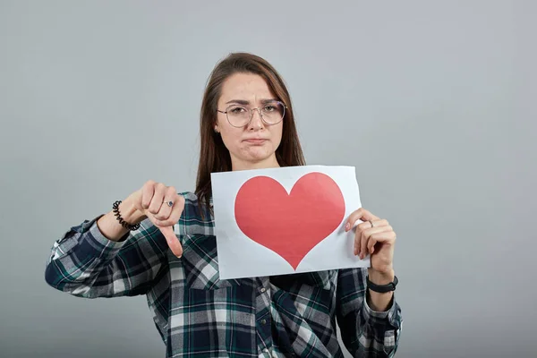 Disappointed woman in glasses holds piece of paper with heart, turned thumb down — Stock Photo, Image