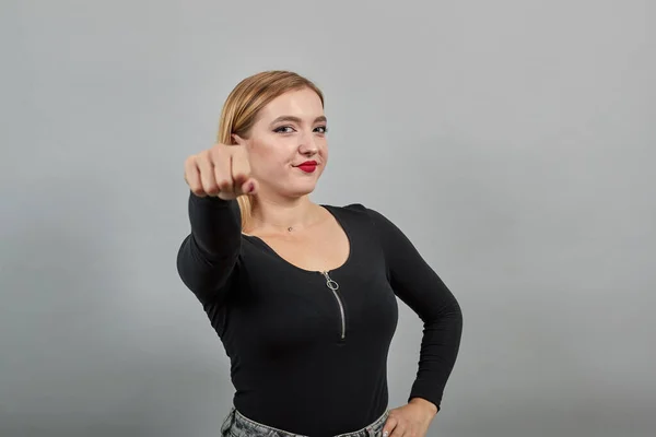 Blonde girl in black jacket on grey background: woman shows off fist, smiles — Stockfoto