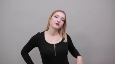 blonde girl in black jacket on grey background: woman shows off fist, smiles