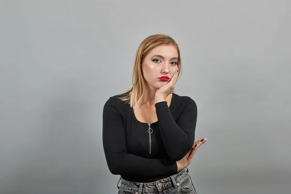 Young blonde girl in black jacket sad woman is bored, touches chin with hand