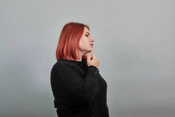 Young redhead fat lady in black sweater woman thinks, touches chin with hand