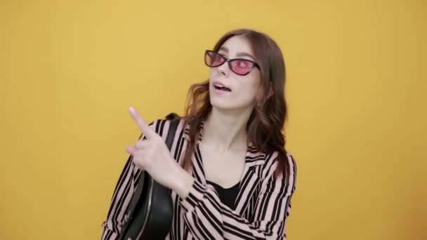 Gelukkig meisje met zonnebril en zak glimlacht, toont richting met wijsvinger — Stockvideo