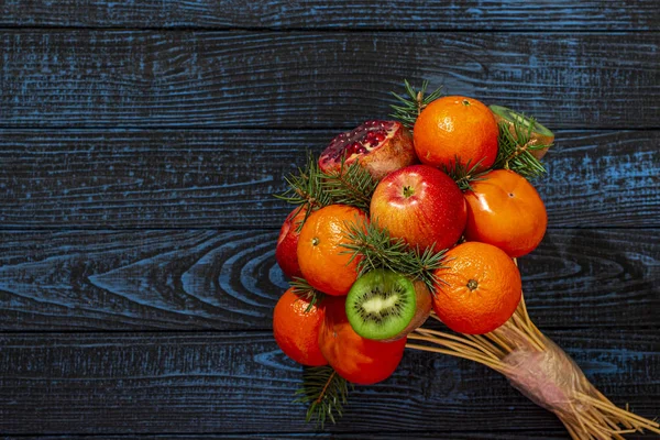 A bouquet of fruits on a blue wooden background consisting of apples, kiwi, pomegranate tangerines and Christmas tree branches.