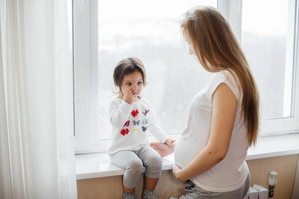 A pregnant mother is standing near the window with her little daughter